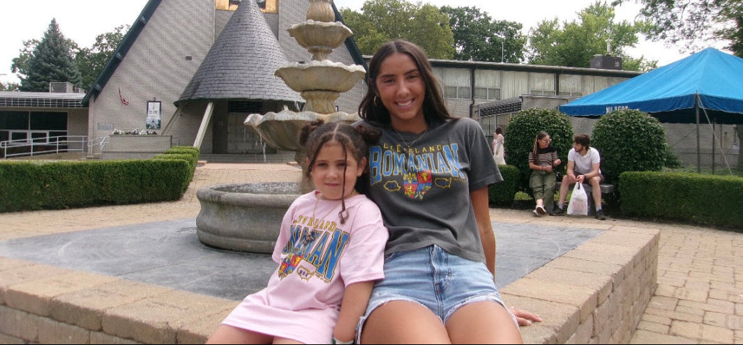 Two young girls sitting on a fountain ledge with a building and trees in the background.